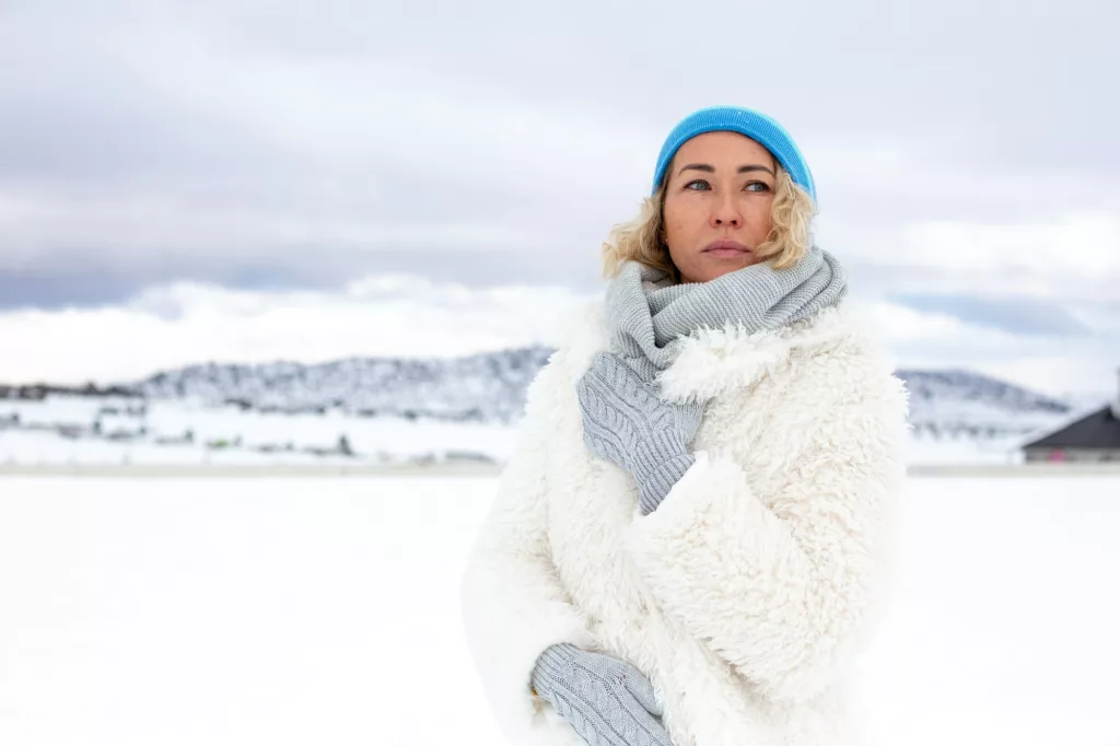 beautiful blonde woman in snowy mountains in blue hat and white eco fir coat. Winter concept