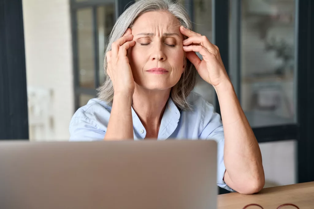 Stressed senior 60s aged massaging head at home office, suffering on workplace.