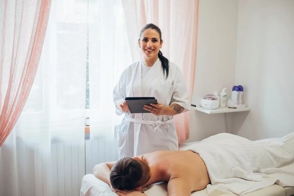 Smiling spa therapist with digital tablet standing by client receiving a massage